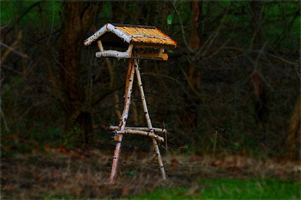 Ein Vogelhaus aus Birkenholz mit Strohdach auf einem klassischen Dreibein-Ständer in einem Park